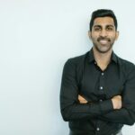 Portrait of a confident man in black shirt smiling against a plain background.